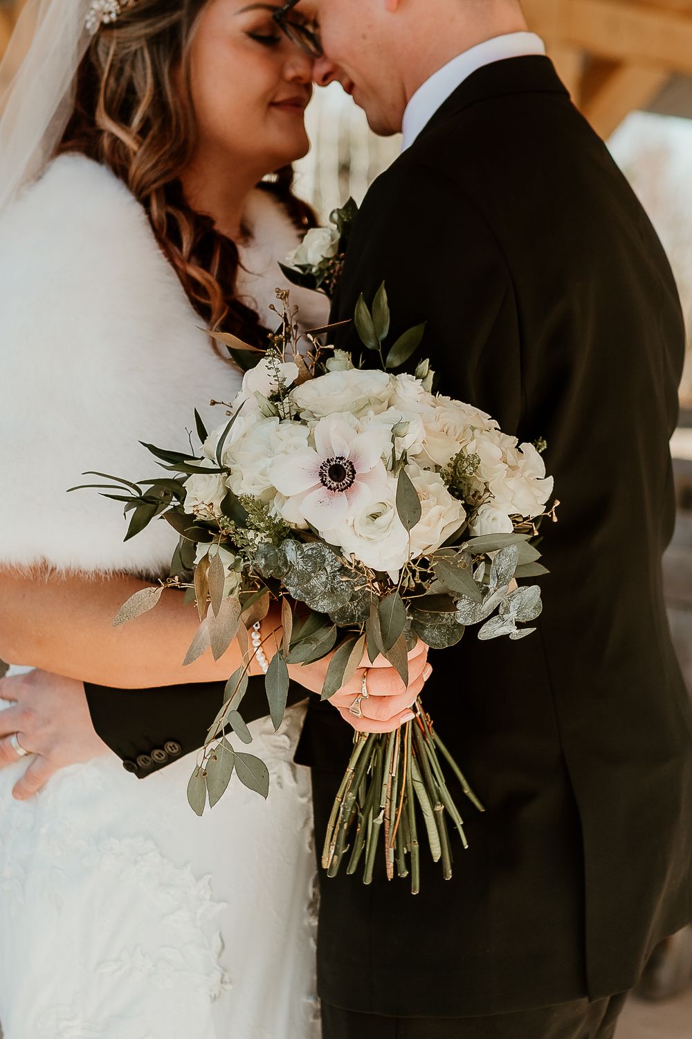Bride and groom share an intimate moment holding a white floral wedding bouquet with greenery and anemones.
