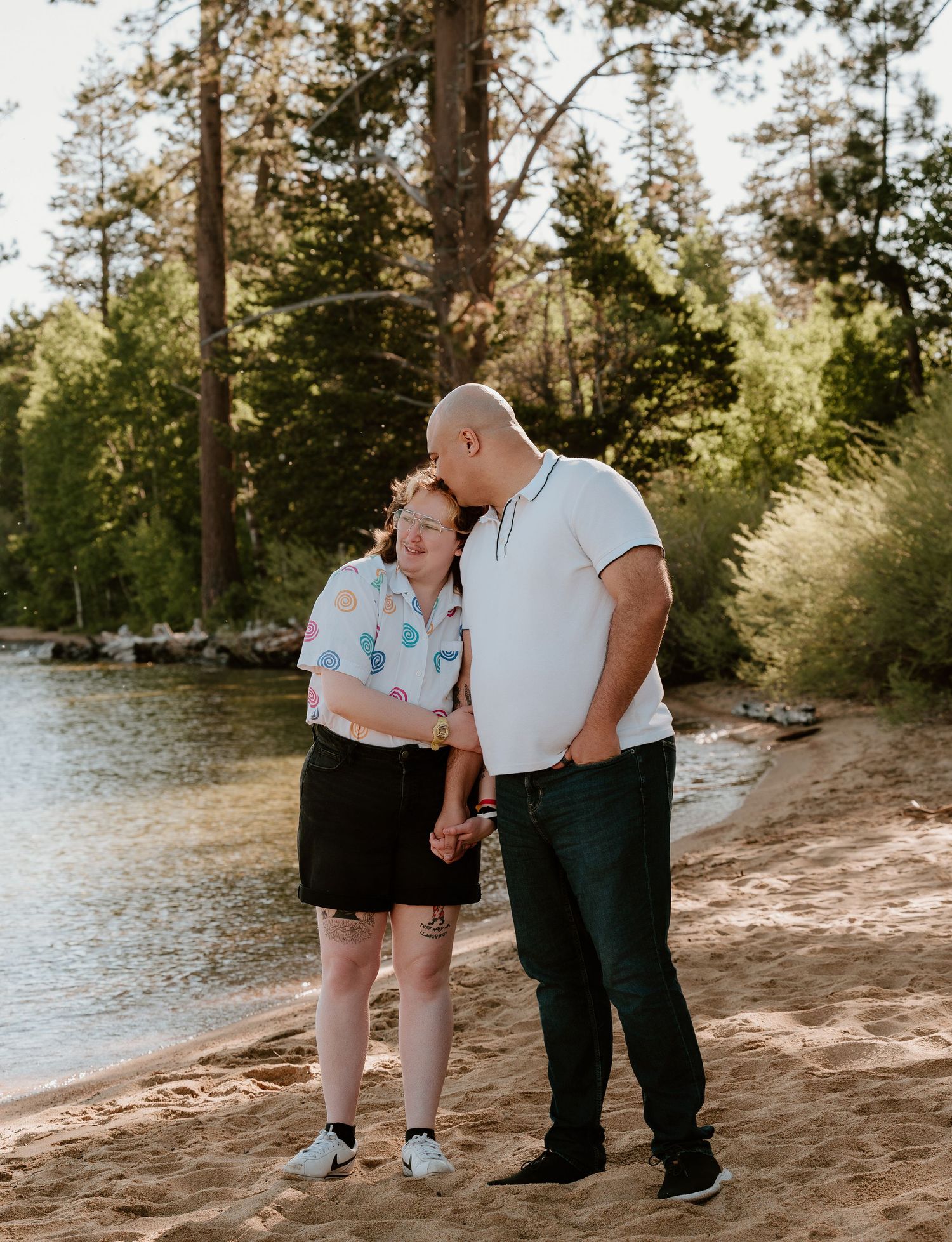 Couple sharing a tender moment on a sandy lakeshore, surrounded by tall pine trees in a serene outdoor setting.