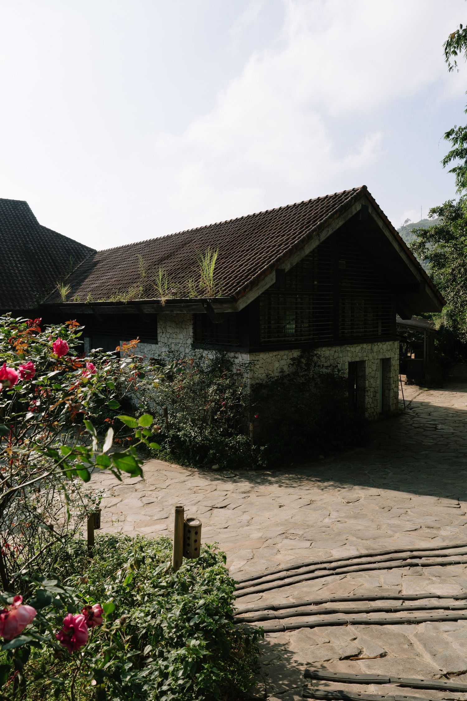 Traditional Asian stone building with tiled roof surrounded by lush greenery, flowering plants, and a cobblestone pathway.