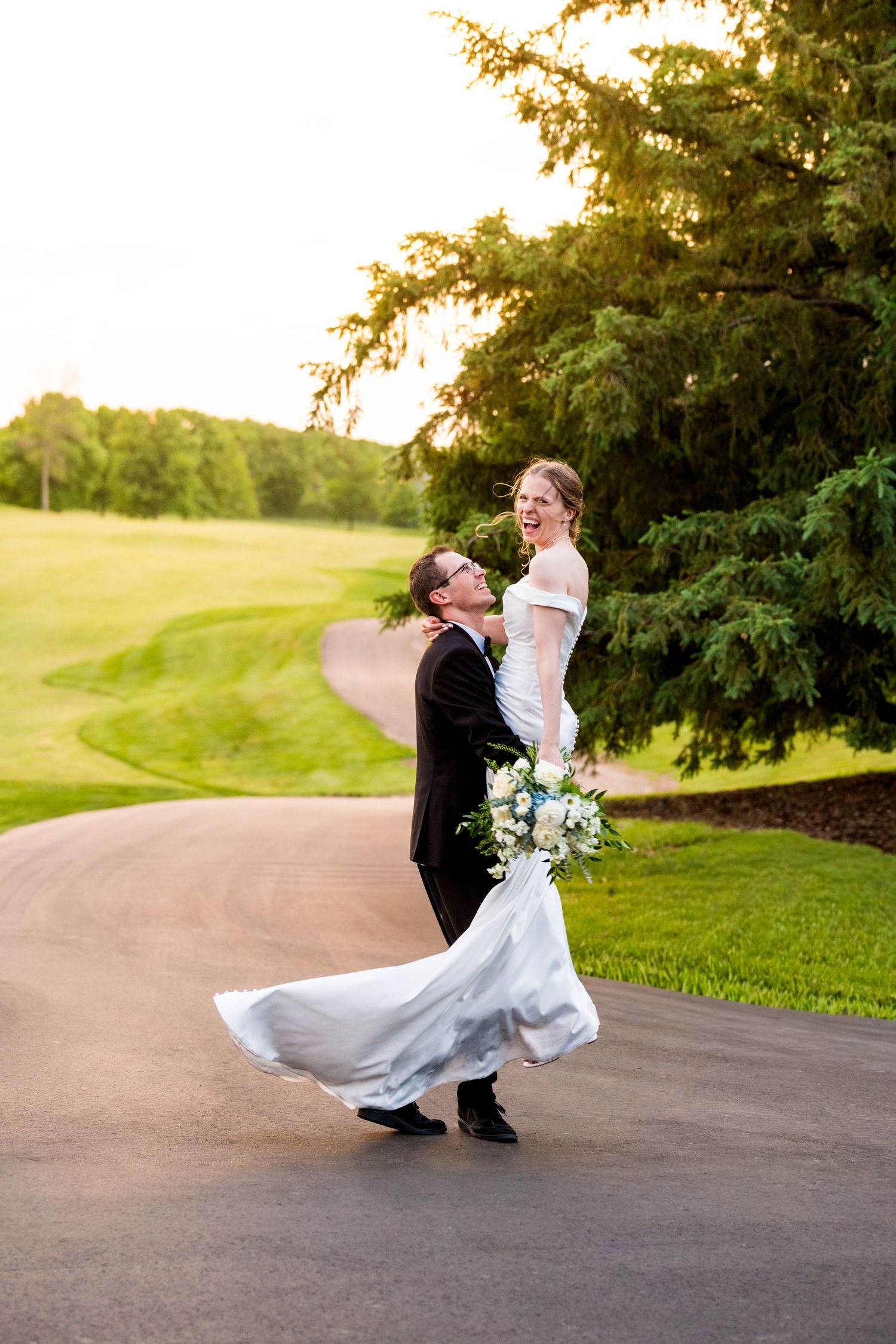 A couple in wedding attire kneels by a white fence to pet horses on a farm with a red barn in the background.