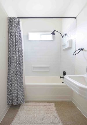 Clean white bathroom with tub-shower combo, gray patterned curtain, wall-mounted fixtures, and small window.
