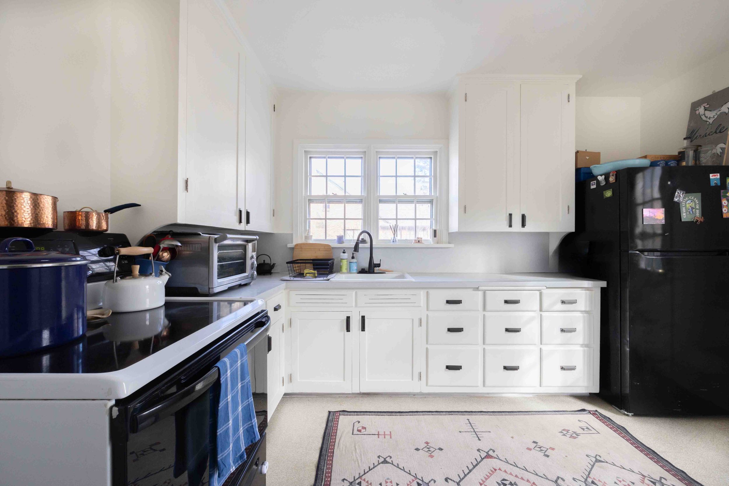 Spacious white kitchen with black appliances, copper pots, decorative rug, and natural window light.