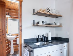 Compact kitchenette with gray countertop, floating shelves, and view into log cabin bathroom.