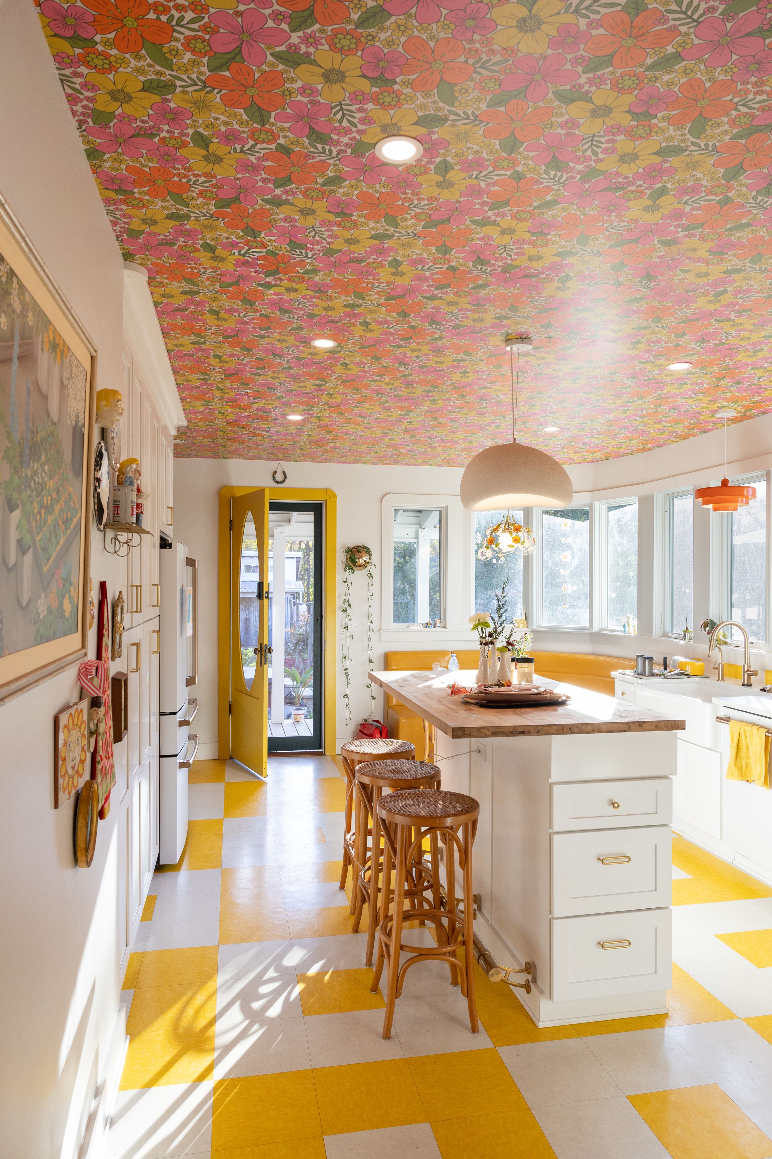 Bright retro kitchen with bold floral wallpapered ceiling, yellow checkered floors, white cabinets, and bar stools.