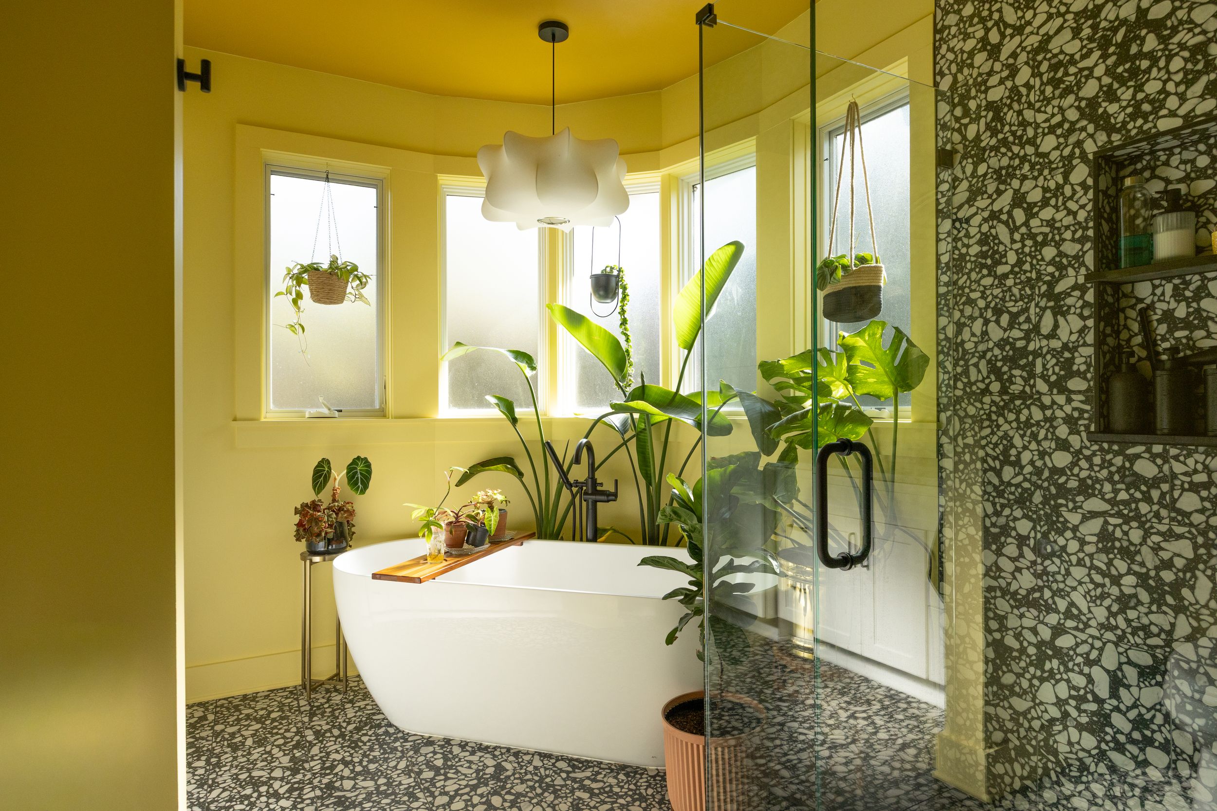 Yellow bathroom with freestanding tub, terrazzo floors, tropical plants, and glass shower enclosure.