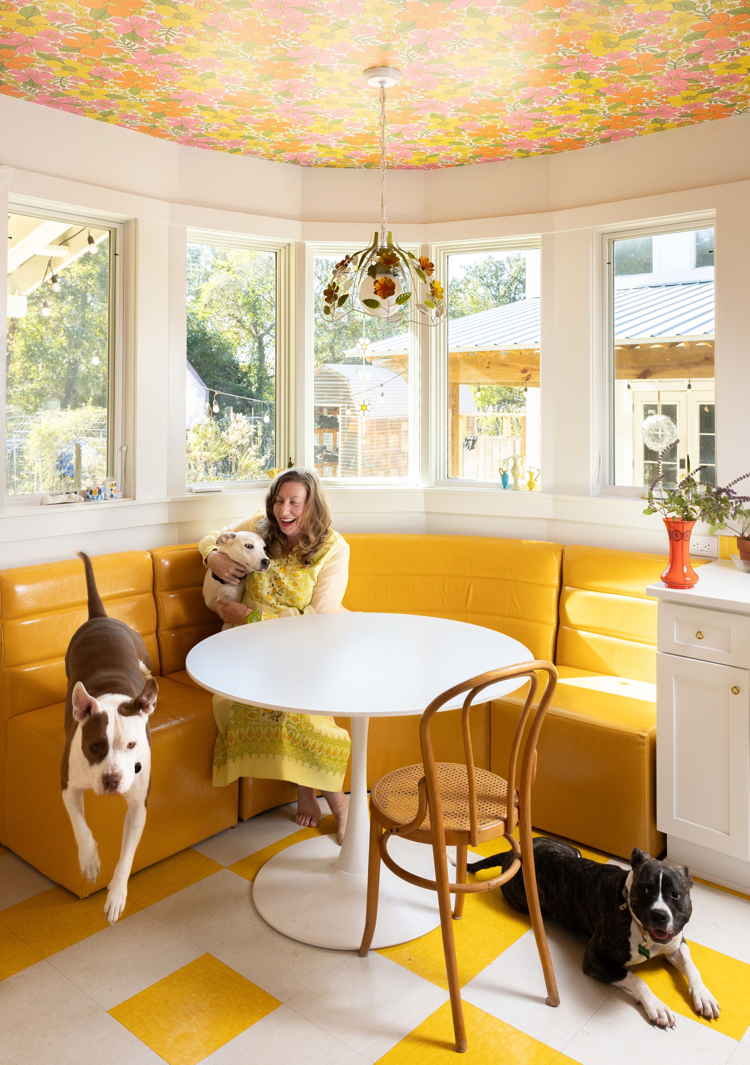 Woman with dogs in yellow banquette breakfast nook with floral ceiling and checkerboard floors.