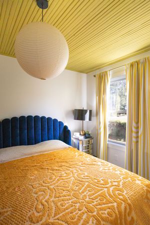 Cozy bedroom with blue velvet headboard, yellow bedspread, and striped curtains.