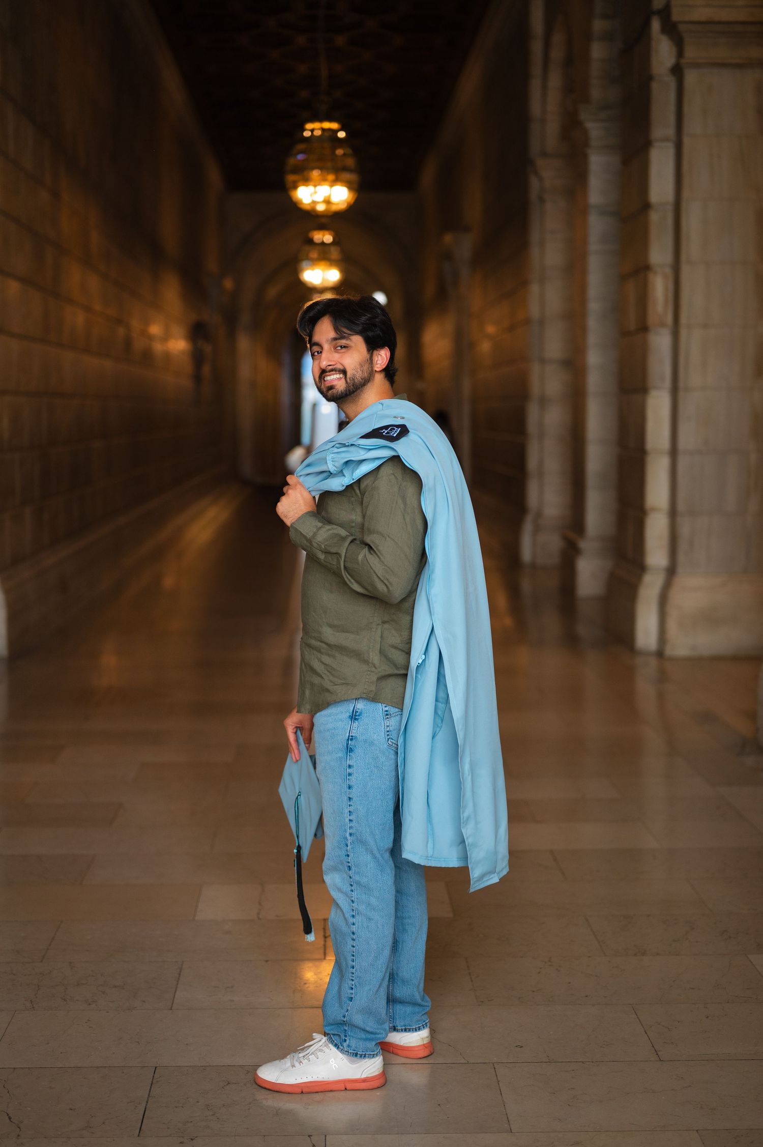 Smiling man in green shirt and jeans holding a light blue jacket in an elegant corridor with arched ceilings.