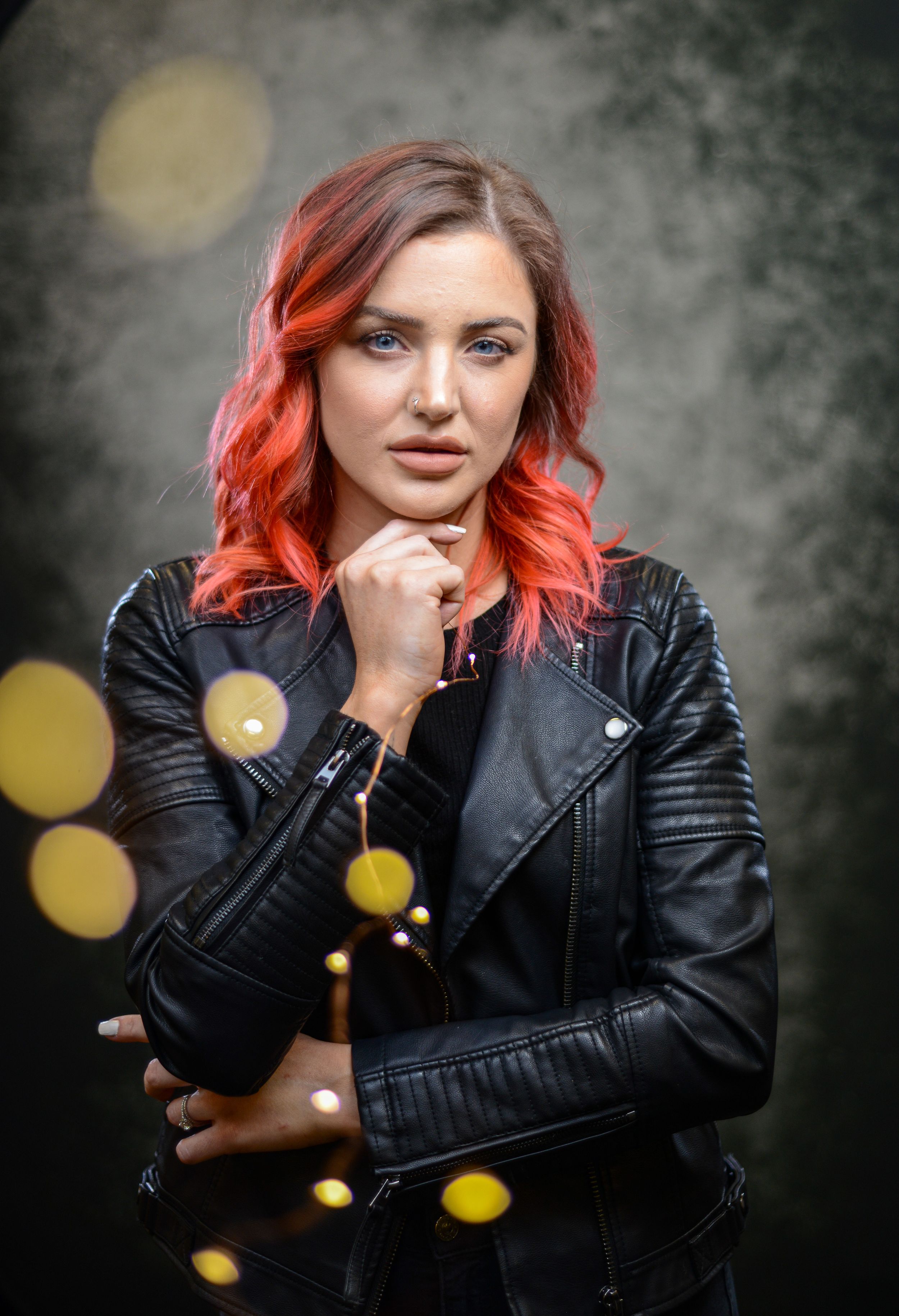 Woman with vibrant red hair wearing a black leather jacket poses confidently against a dark bokeh background.
