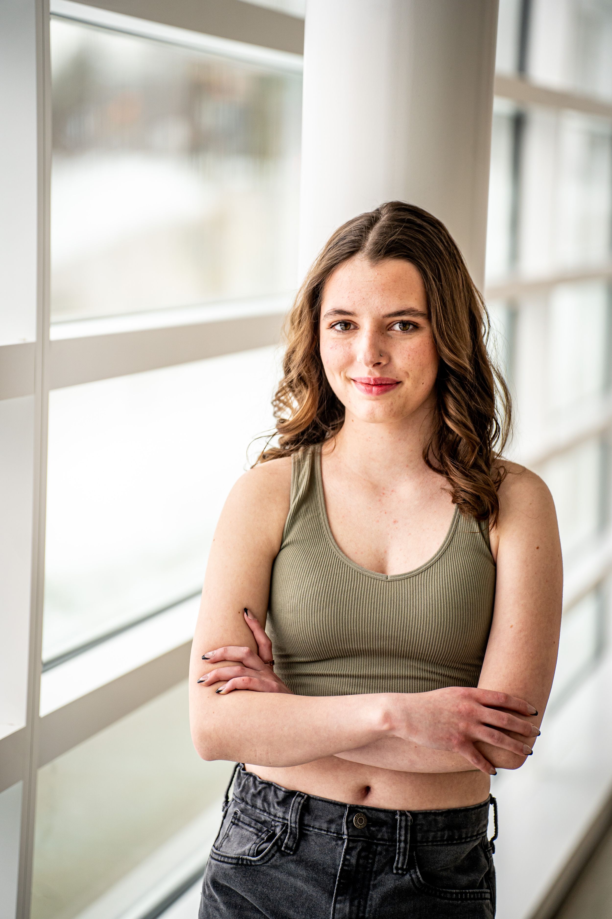 Young woman in olive crop top and jeans stands arms crossed by bright windows, smiling confidently.