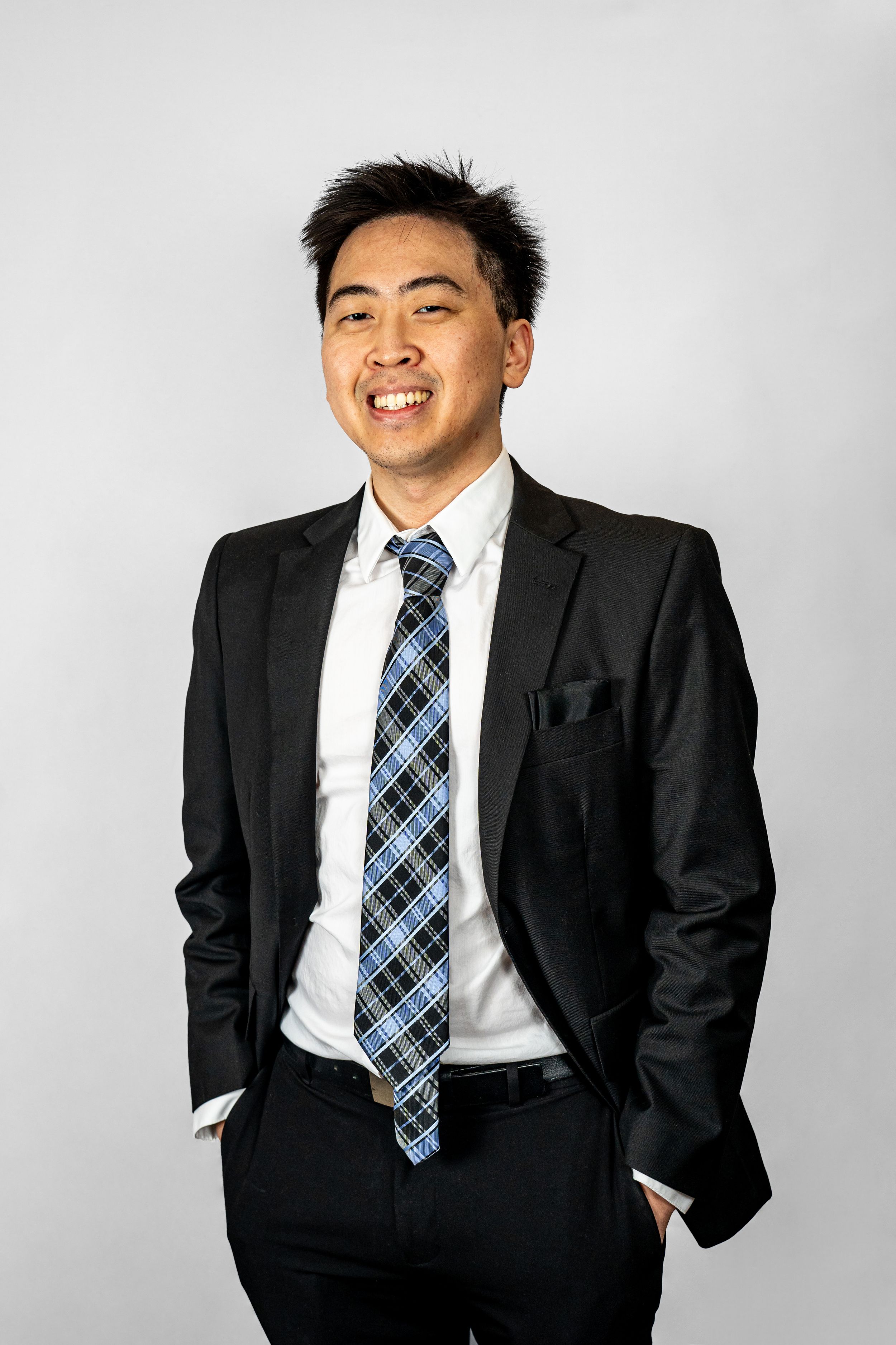 Professional headshot photo of smiling young Asian businessman in black suit and plaid tie posing confidently with hands in pockets.