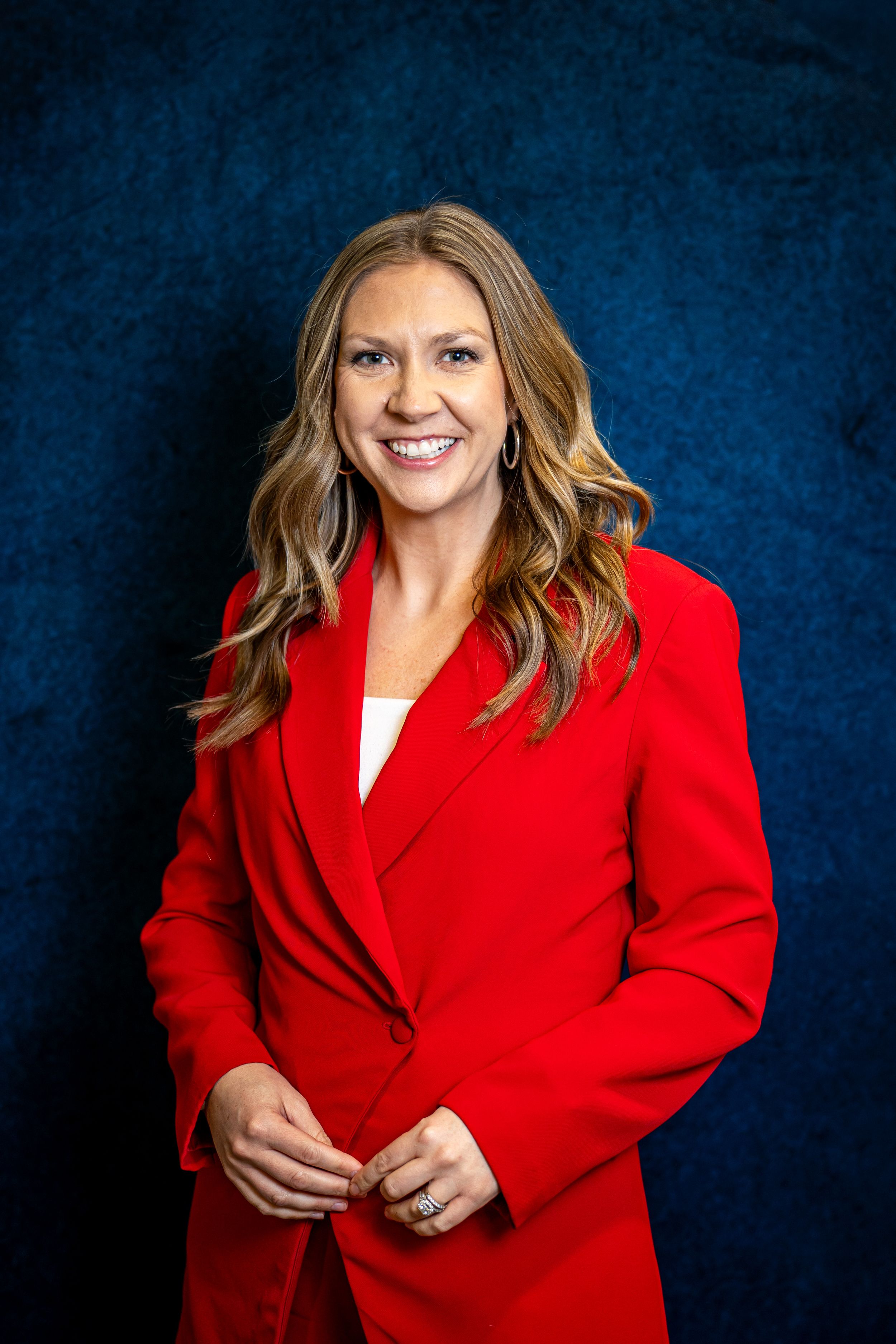 Professional headshot image of a woman in red blazer smiling confidently against dark blue background, business portrait photography.