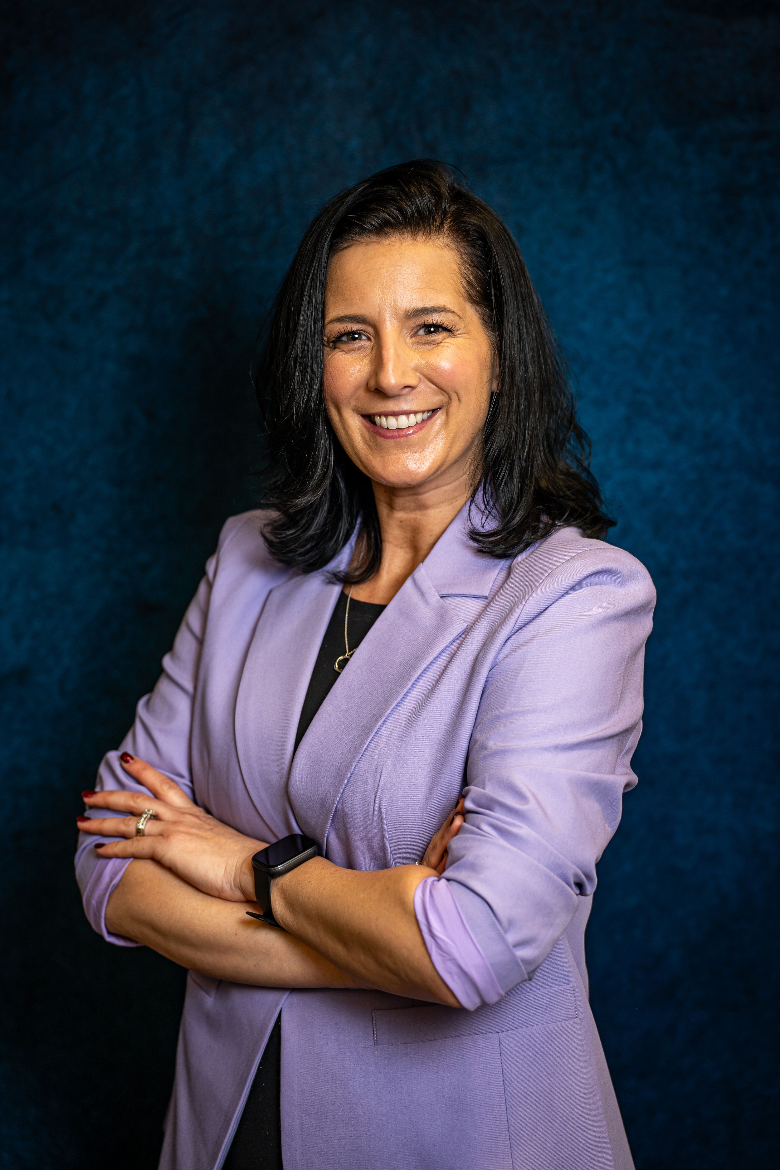 Professional woman in lavender blazer smiling with arms crossed against dark blue background. corporate headshot