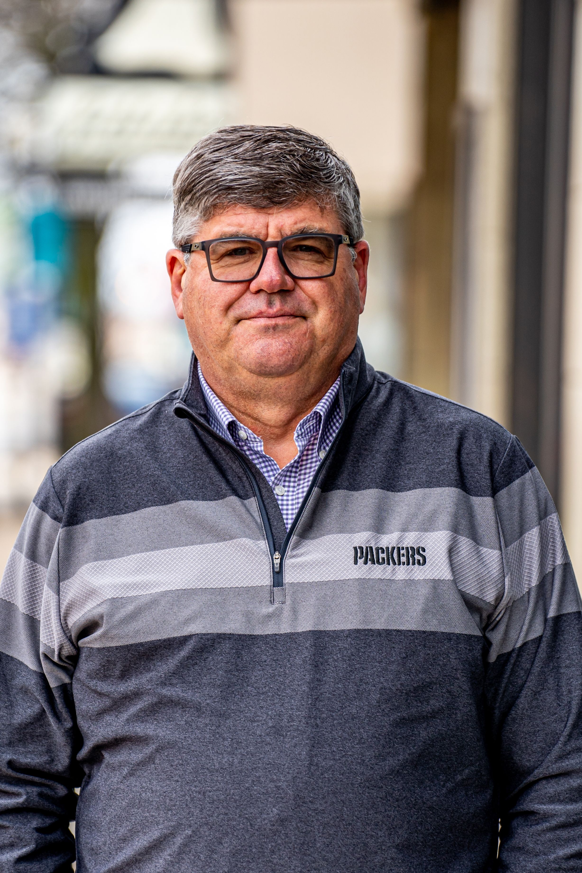 Middle-aged man wearing Green Bay Packers quarter-zip pullover and glasses standing outdoors on a city street.