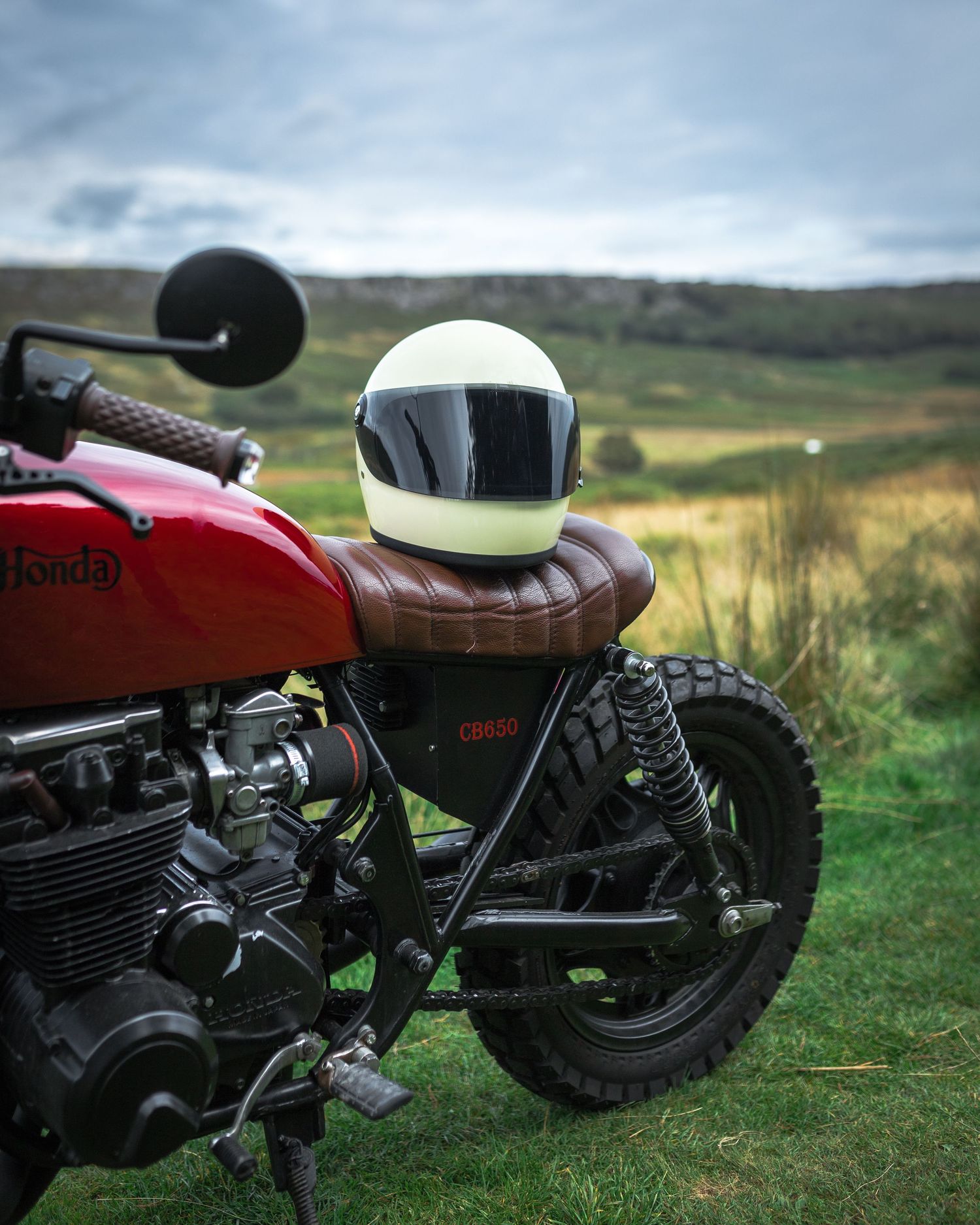 Red Honda CB650 cafe racer motorcycle with white helmet on seat, parked in scenic green countryside under cloudy sky.