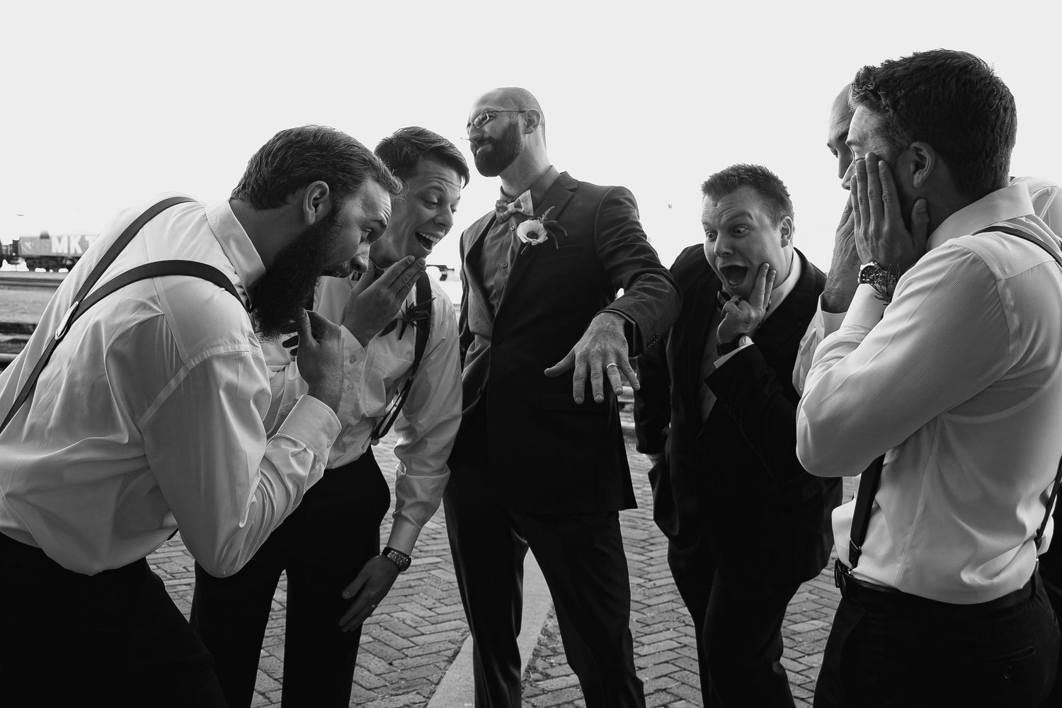 Black and white photo of a groom in a suit laughing with groomsmen in suspenders at a wedding.