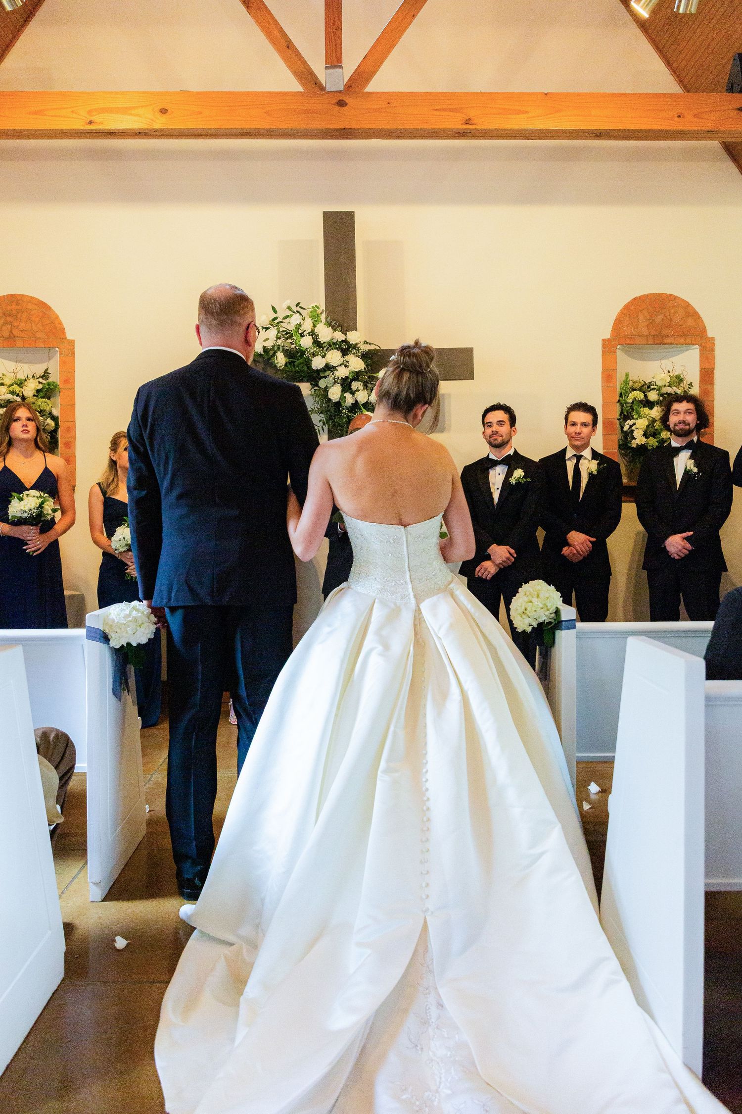Bride in strapless white ball gown stands at church altar with groom and groomsmen in black tuxedos during wedding ceremony.