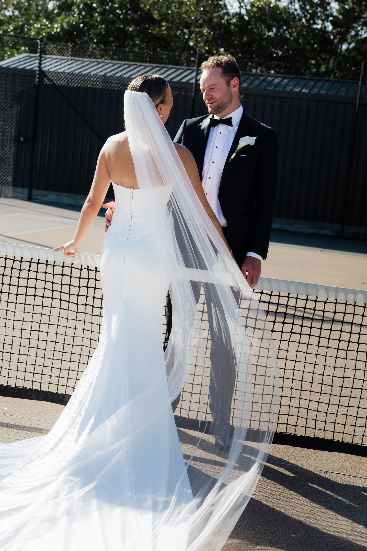 Bride in white mermaid gown with flowing veil and groom in tuxedo posing by tennis net on wedding day.