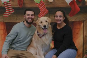 Smiling couple poses with their golden retriever dog in front of a festive Christmas fireplace backdrop.