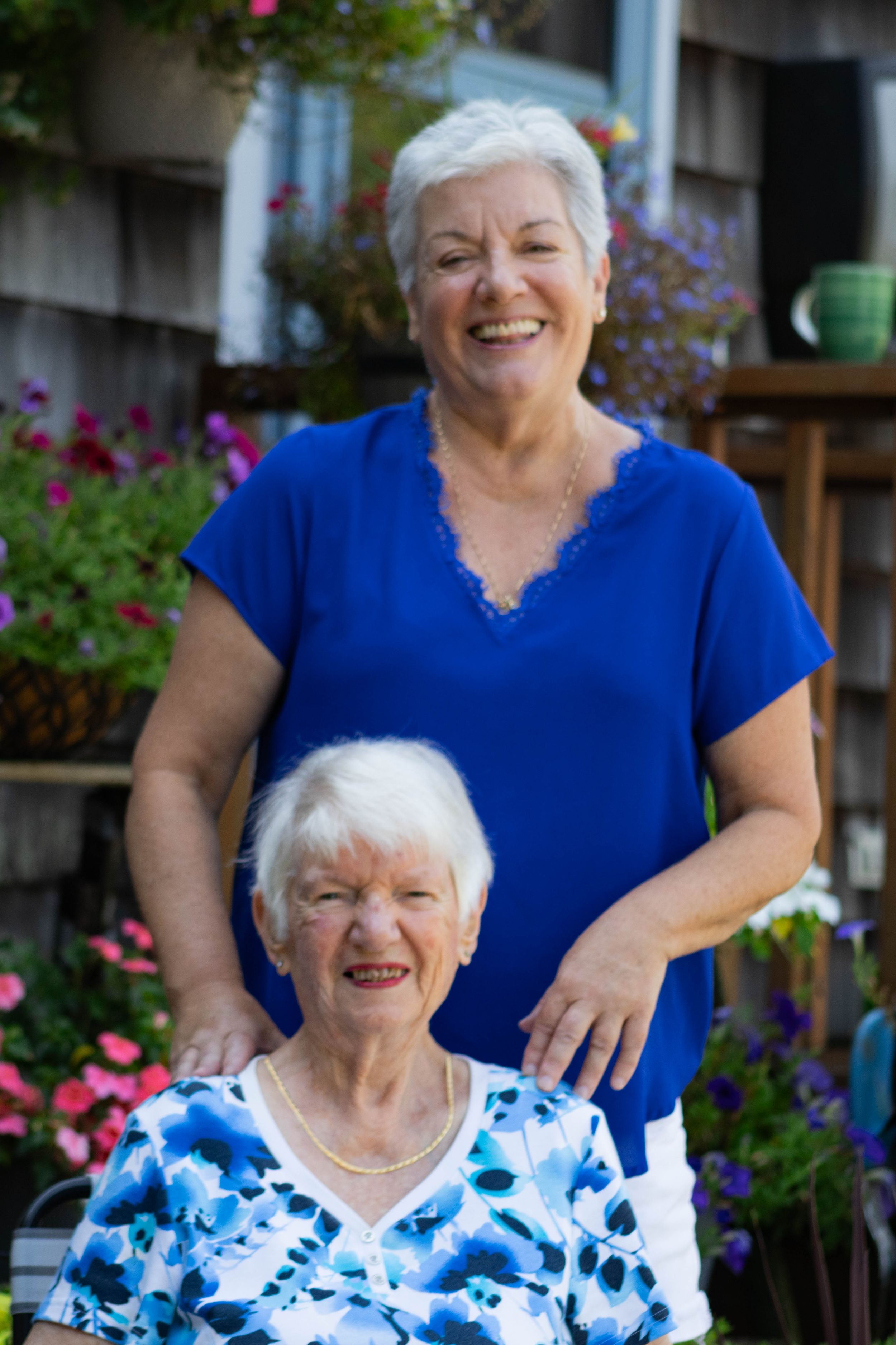 Two women, mother and daughter, smiling outdoors near a flower-filled garden, one seated and one standing.