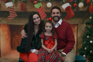 Family of three with young daughter in red dress poses before a decorative Christmas fireplace backdrop.