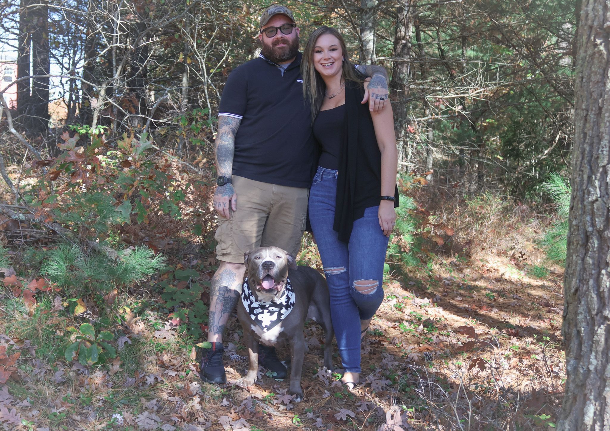 Couple stands with their pit bull dog in a wooded autumn forest setting, dog wearing bandana.
