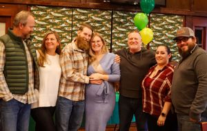 Group of seven adults posing together at a festive green and gold balloon decorated event.