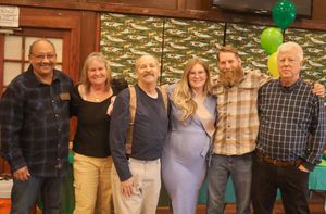 Six adults posing together at a party with green balloon and airplane themed decorations.
