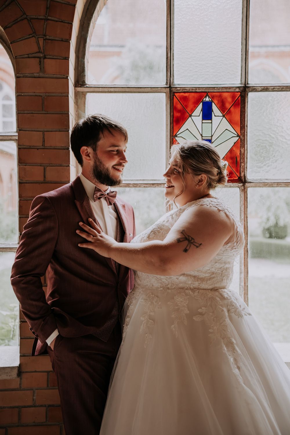 Bride and groom share a romantic moment by a stained glass window, bride in lace gown, groom in burgundy suit.