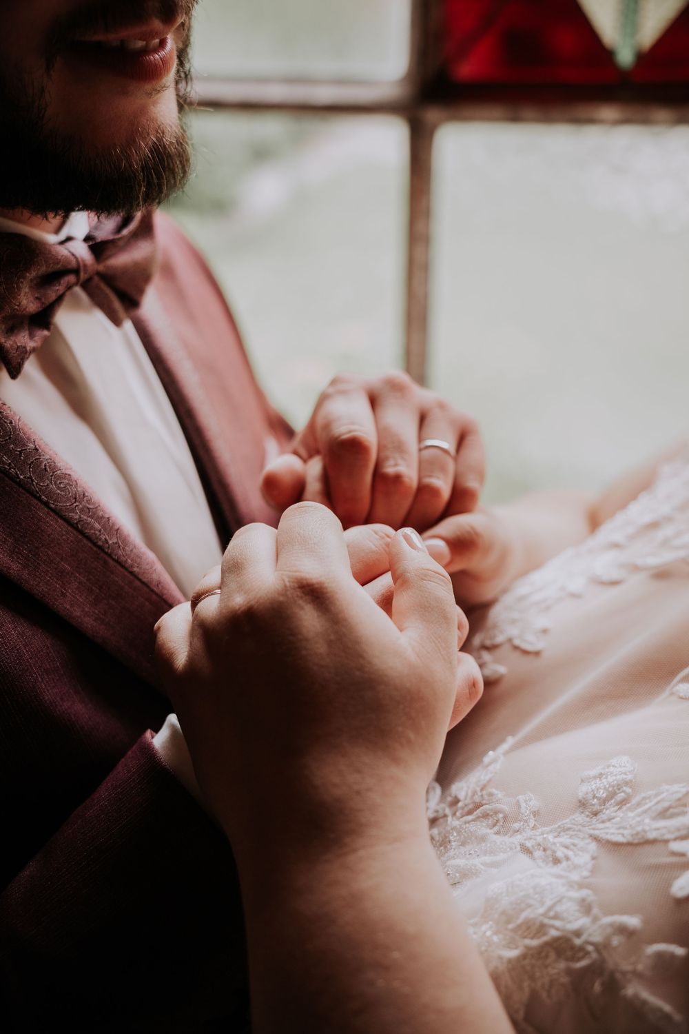 Close-up of bride and groom holding hands with wedding rings visible, groom wearing a burgundy suit and bow tie.