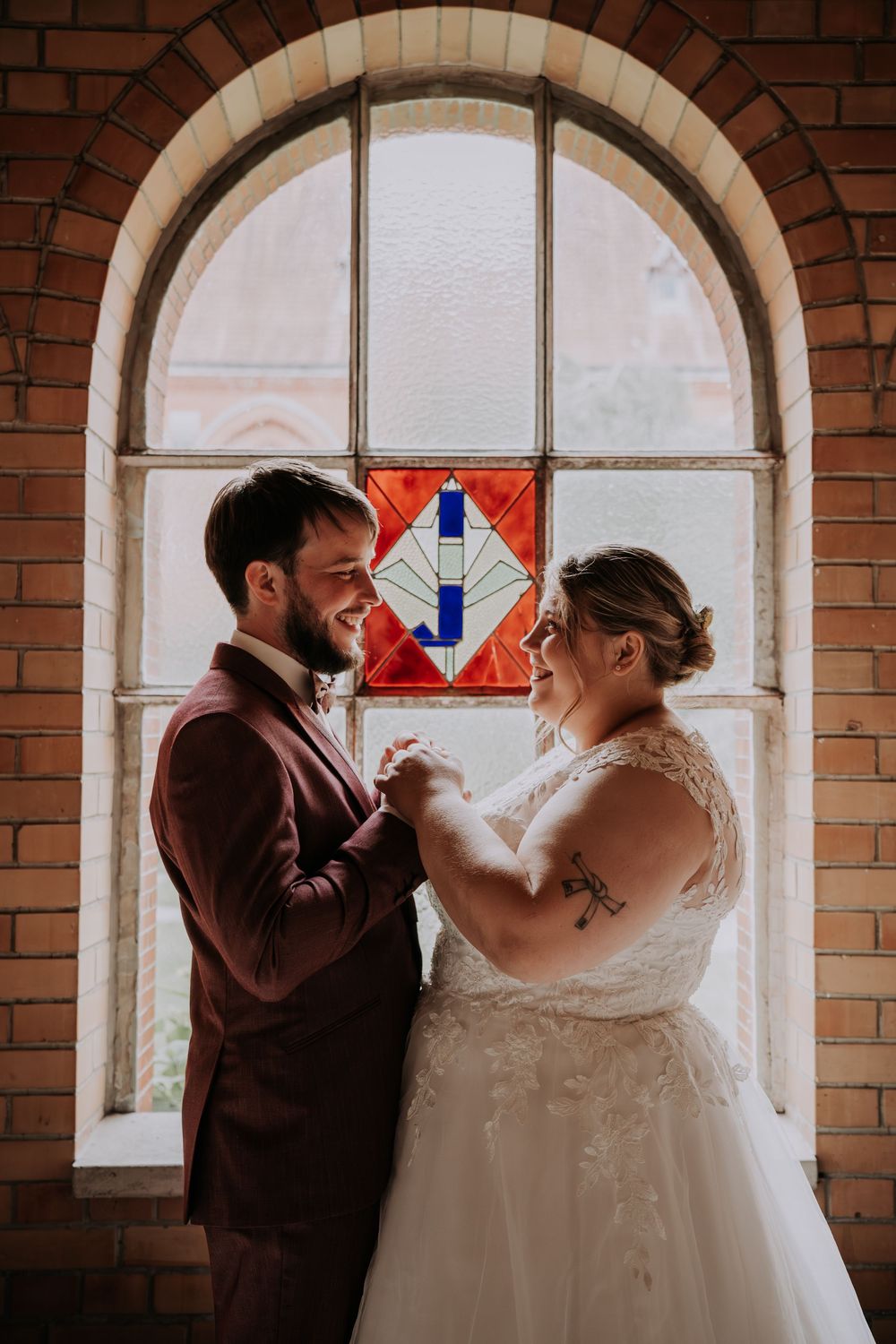 Bride and groom smiling at each other holding hands in front of a stained glass arched window in a brick church.