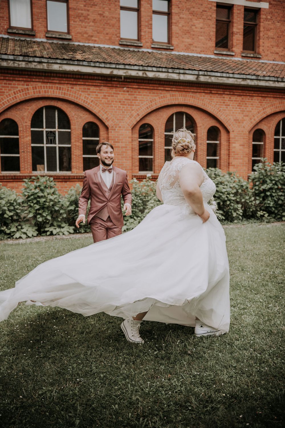 Bride in flowing white gown twirls toward smiling groom in pink suit on lawn before red brick building.
