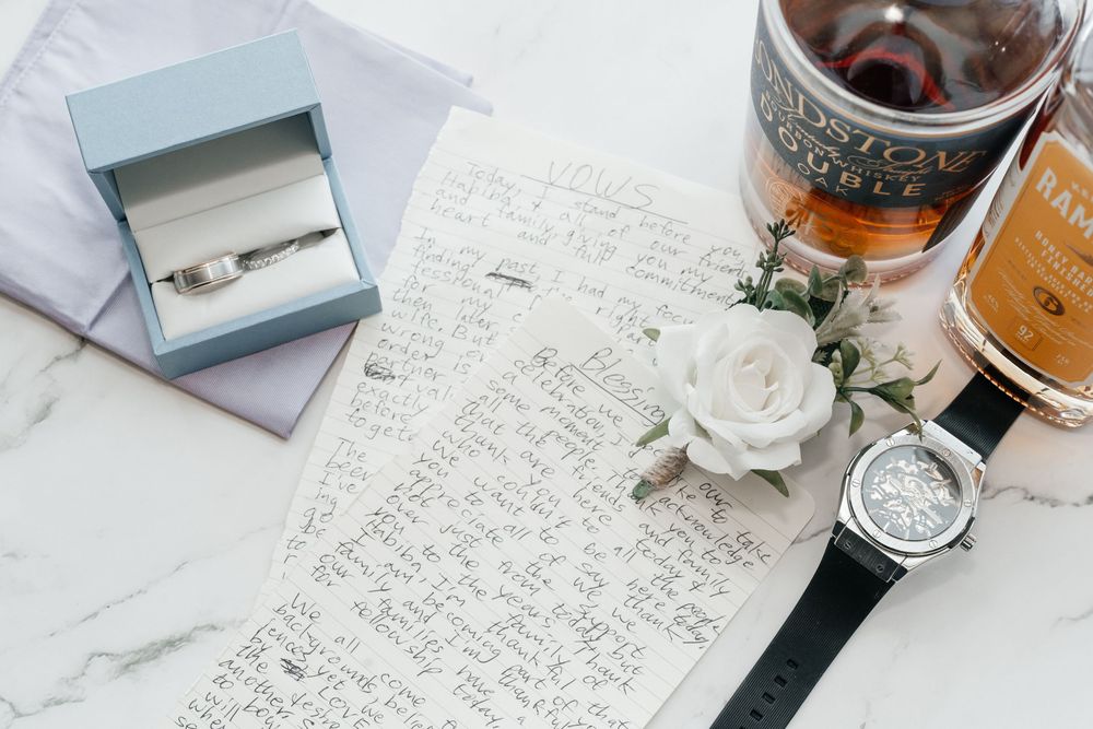 Wedding flat lay with diamond ring in blue box, handwritten vows, white rose, watch, and whiskey glass on marble.