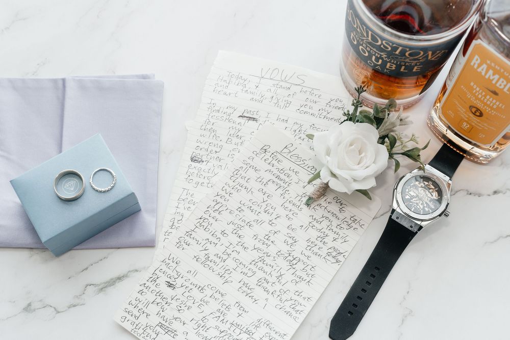 Wedding flat lay with rings, handwritten vows, watch, boutonniere, and whiskey bottle on marble.