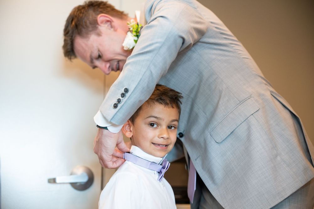 A man in a gray suit adjusts a young boy's tie at a wedding ceremony.