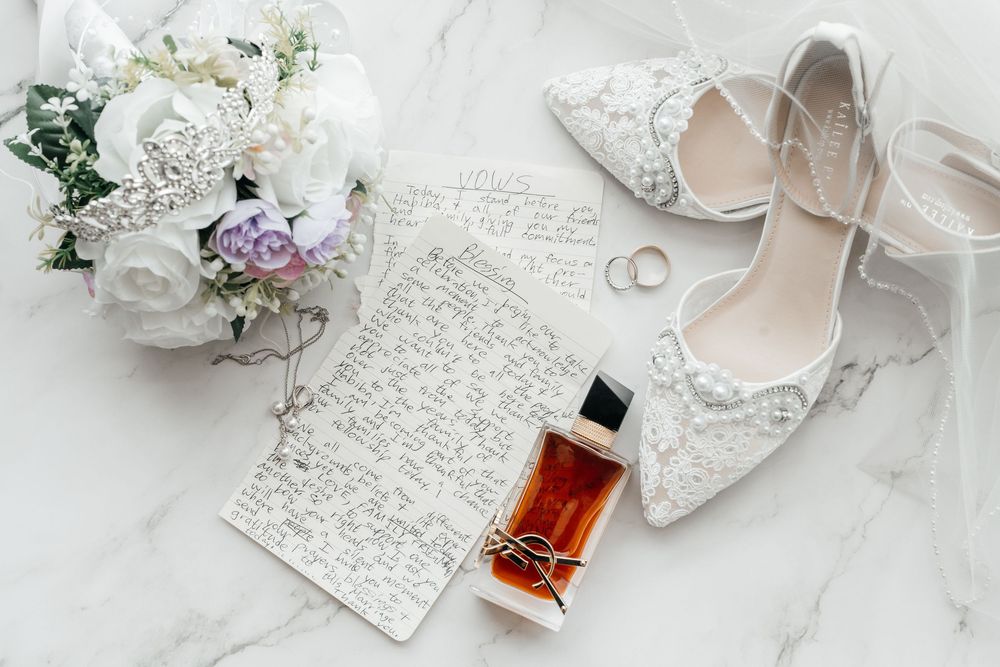 Elegant wedding flat lay with lace heels, bouquet, perfume, rings, and handwritten vows on marble.