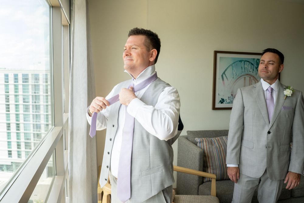 Groom adjusting his lavender tie in a hotel room while groomsman watches, both wearing light gray wedding suits.