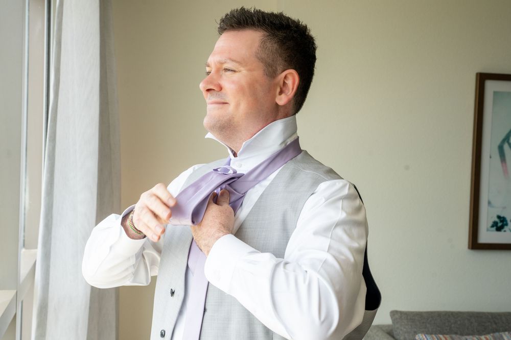 Groom in gray vest adjusting lavender tie near window, preparing for wedding ceremony.