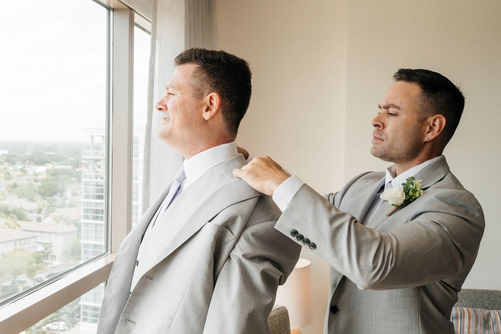 Groomsman helping groom adjust his gray suit jacket near a large window on wedding day.