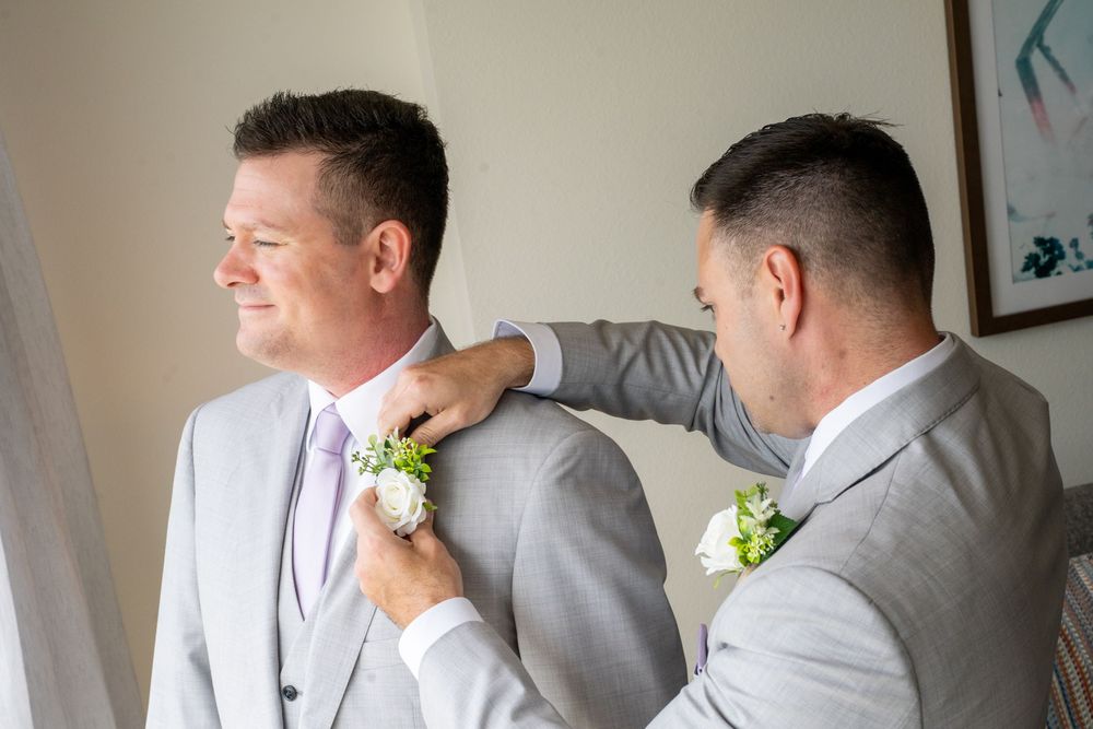 Two men in gray suits prepare for a wedding as one pins a white boutonniere on the other's lapel.