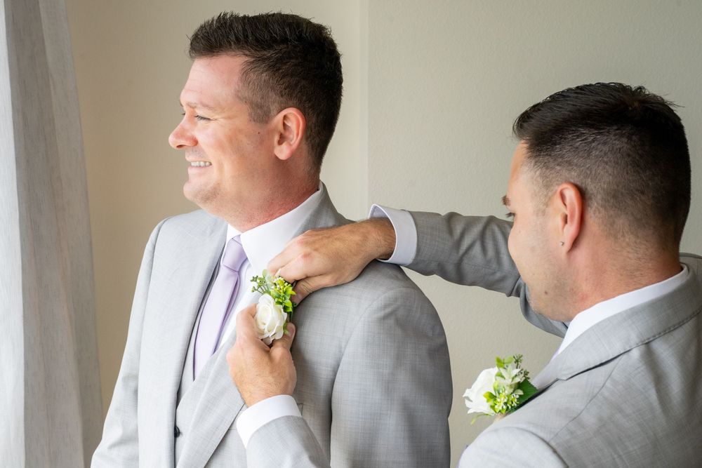 Two grooms in gray suits pinning boutonniere before wedding ceremony.