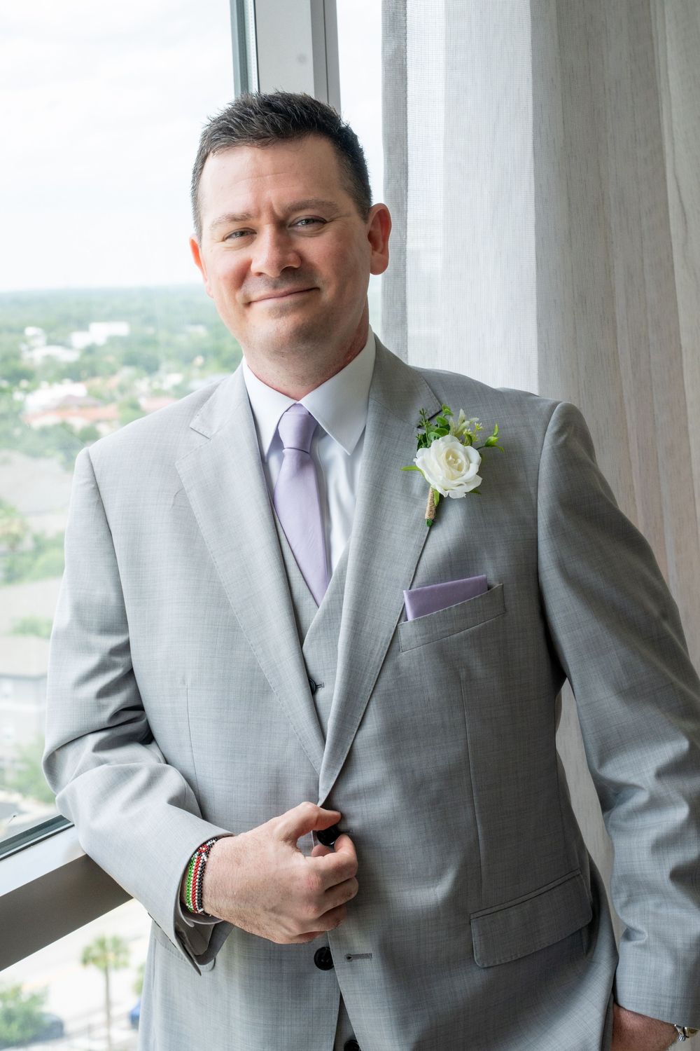 A smiling groom in a light gray suit with lavender tie and white rose boutonniere poses by a window.