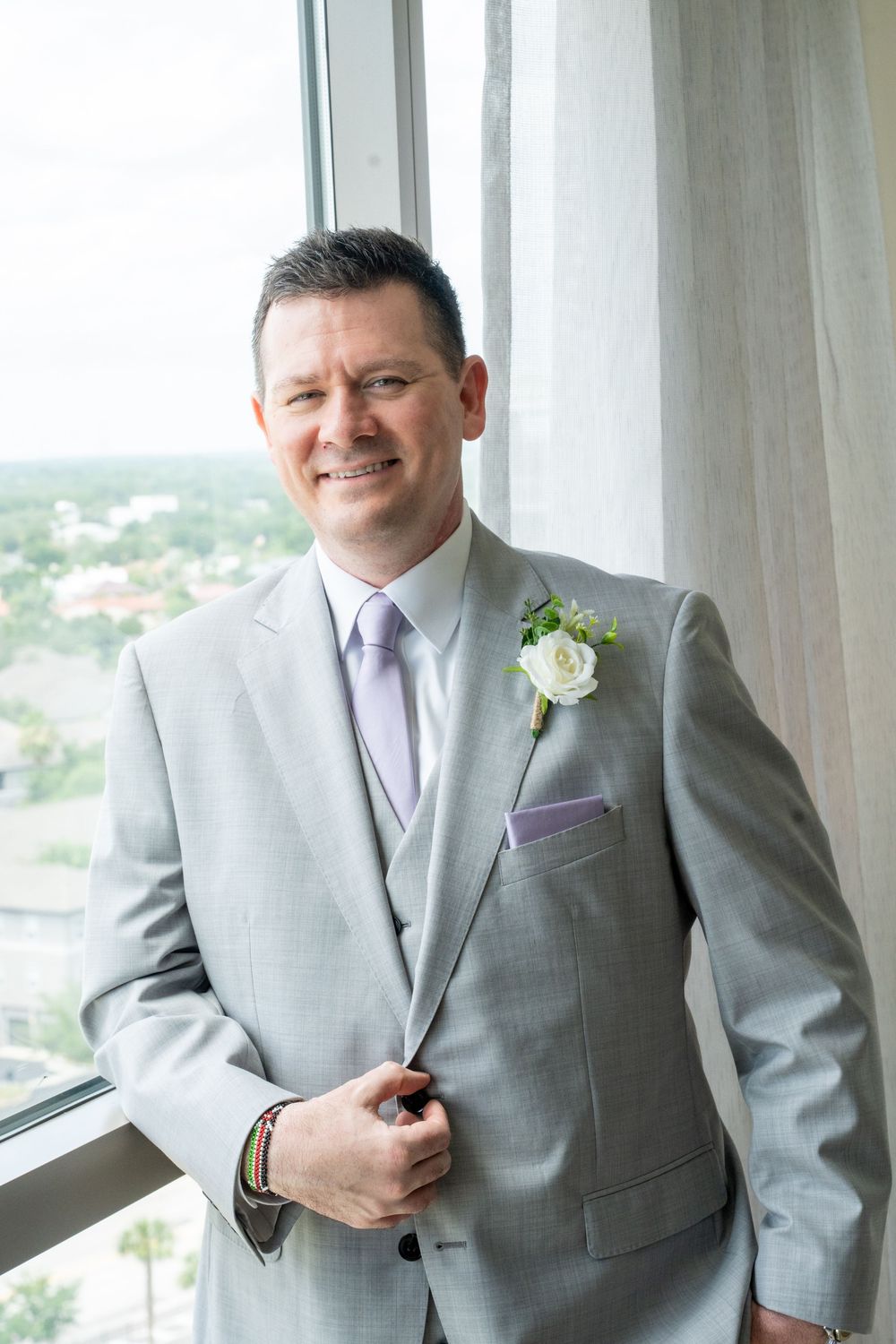 Smiling groom in light gray suit with white rose boutonniere and lavender tie standing by large window.
