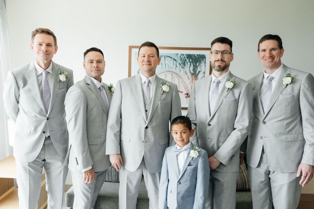 Groom and groomsmen in matching light gray suits with white boutonnieres pose with young ring bearer in blue suit.