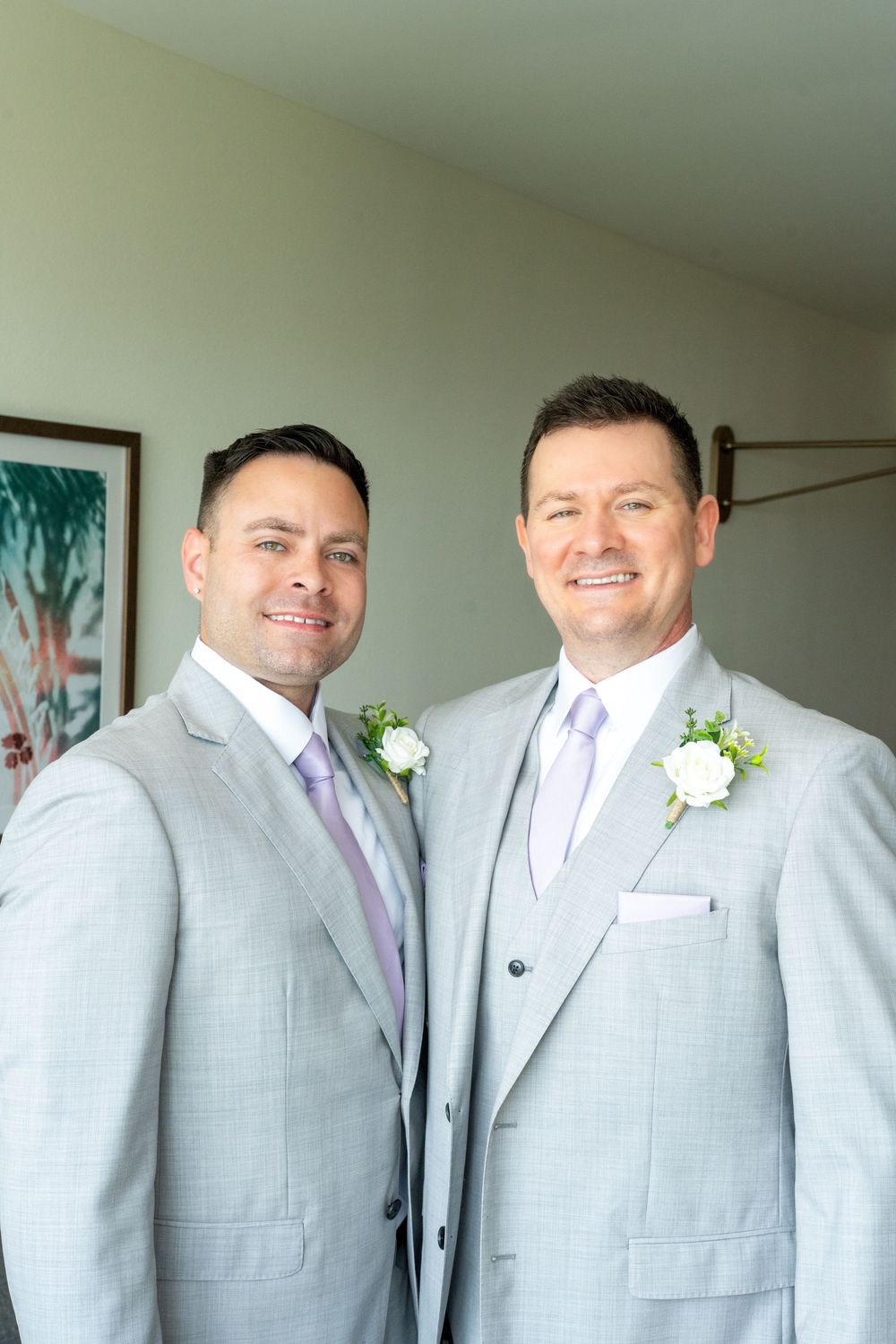 Two grooms in matching light gray suits with lavender ties and white boutonnieres smile together on their wedding day.