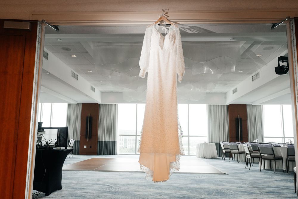 Elegant white long-sleeve wedding dress hanging on a hanger in a bright hotel ballroom with large windows.