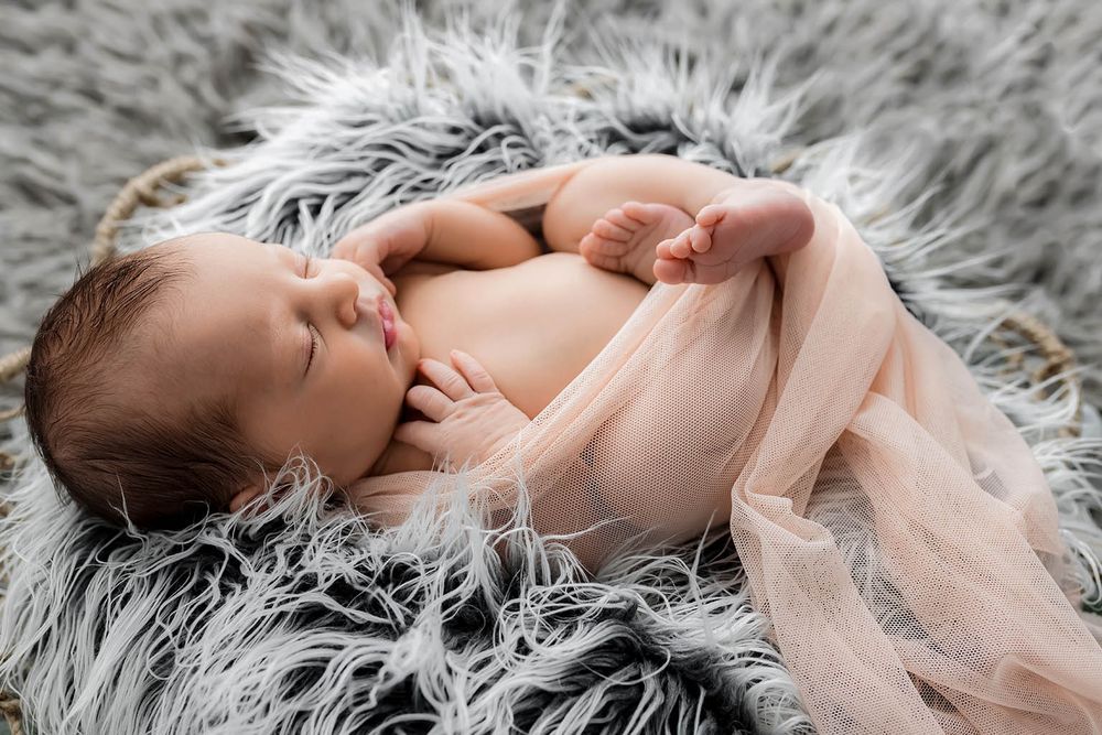 Newborn baby sleeping peacefully on a fluffy gray fur blanket wrapped in a soft pink tulle fabric.
