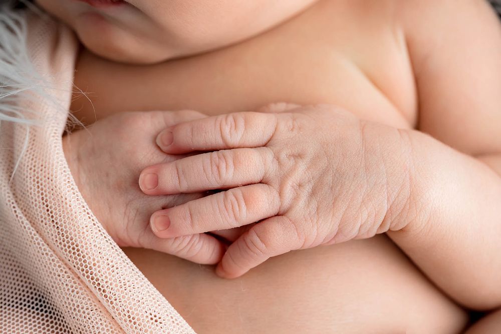 Close-up of a newborn baby's tiny wrinkled hands resting on their chest, with soft pink fabric in background.