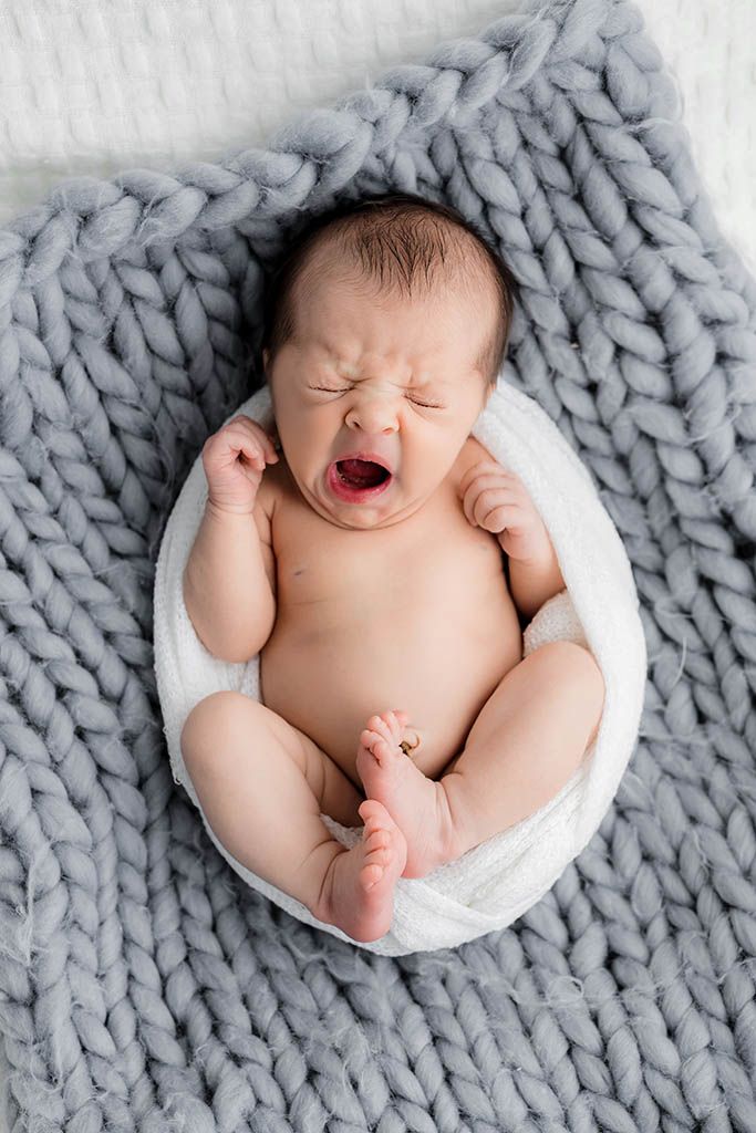 Newborn baby yawning while lying in a white wrap on a chunky gray knit blanket during a studio photo session.