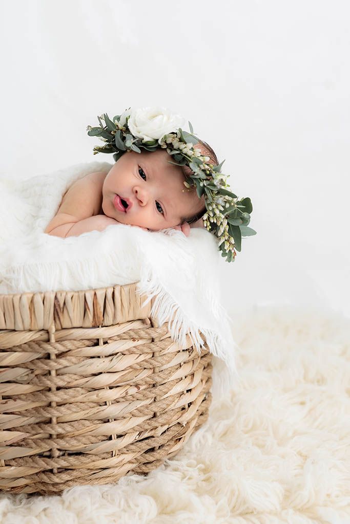 Newborn baby wearing a floral crown resting in a wicker basket lined with white fur blanket.